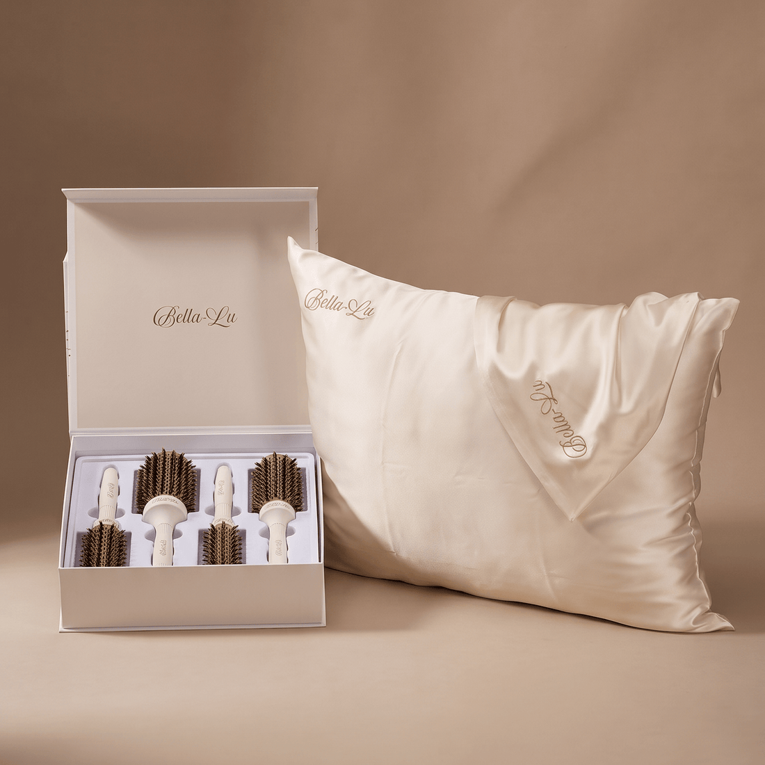 An open hair brush set displaying 4 Bella-Lu round brushes next to a cream silk pillow with gold Bella-Lu embroidery, set against a warm beige studio backdrop.
